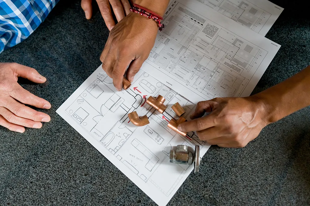 Hands reviewing copper plumbing fittings on a blueprint, discussing pipe installation for HVAC services in British Colum